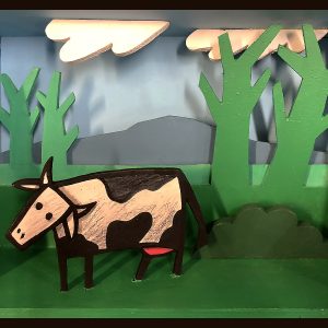 Diorama représentant une vache stylisée dans un décor végétal simplifié avec arbres et collines, en acrylique sur bois