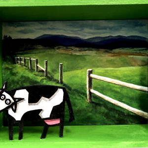 Diorama représentant une vache devant un paysage de prairie avec clôture et collines, en acrylique sur bois